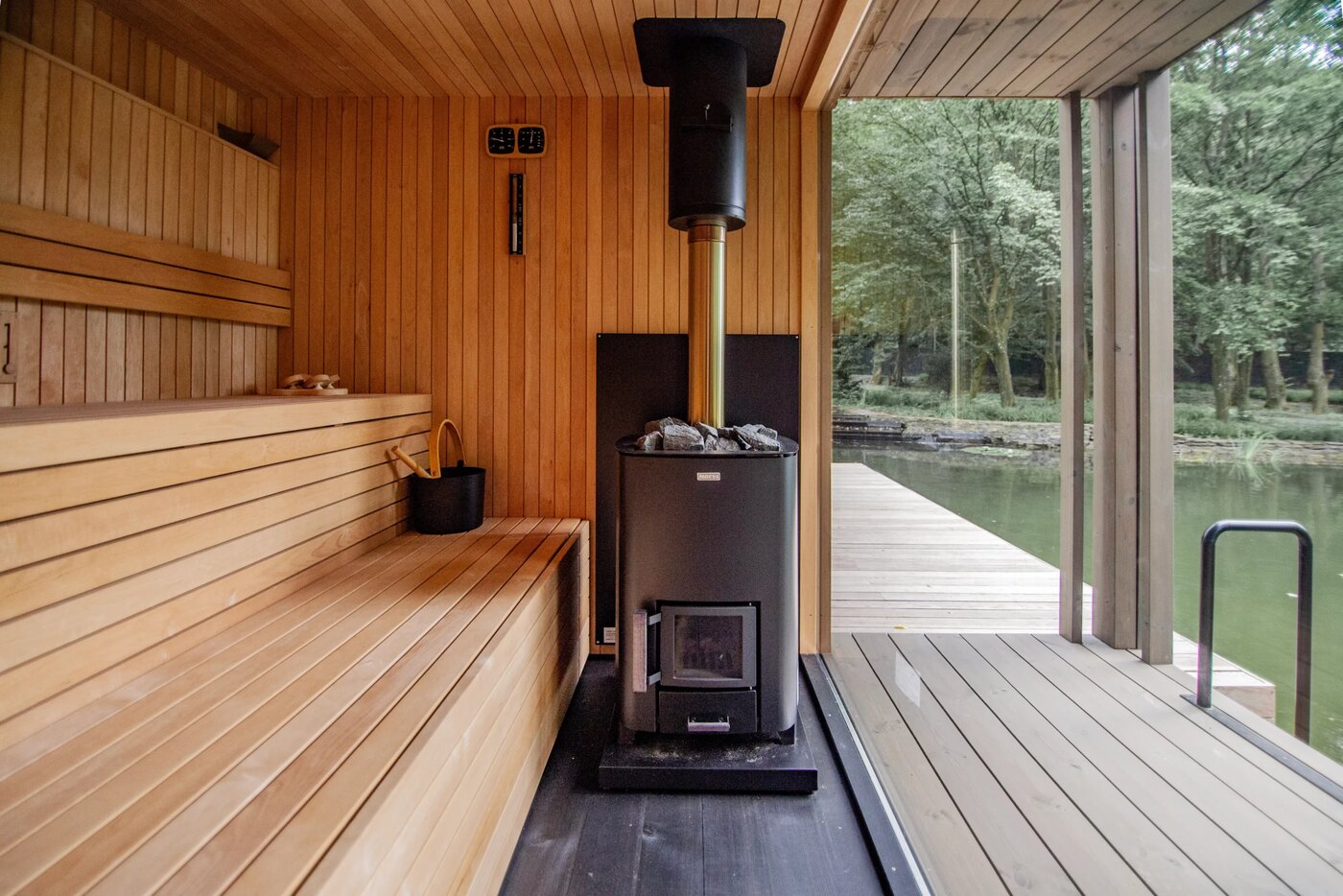 Standalone outdoor sauna opening onto a deck and wooded waterfront through a full glass wall